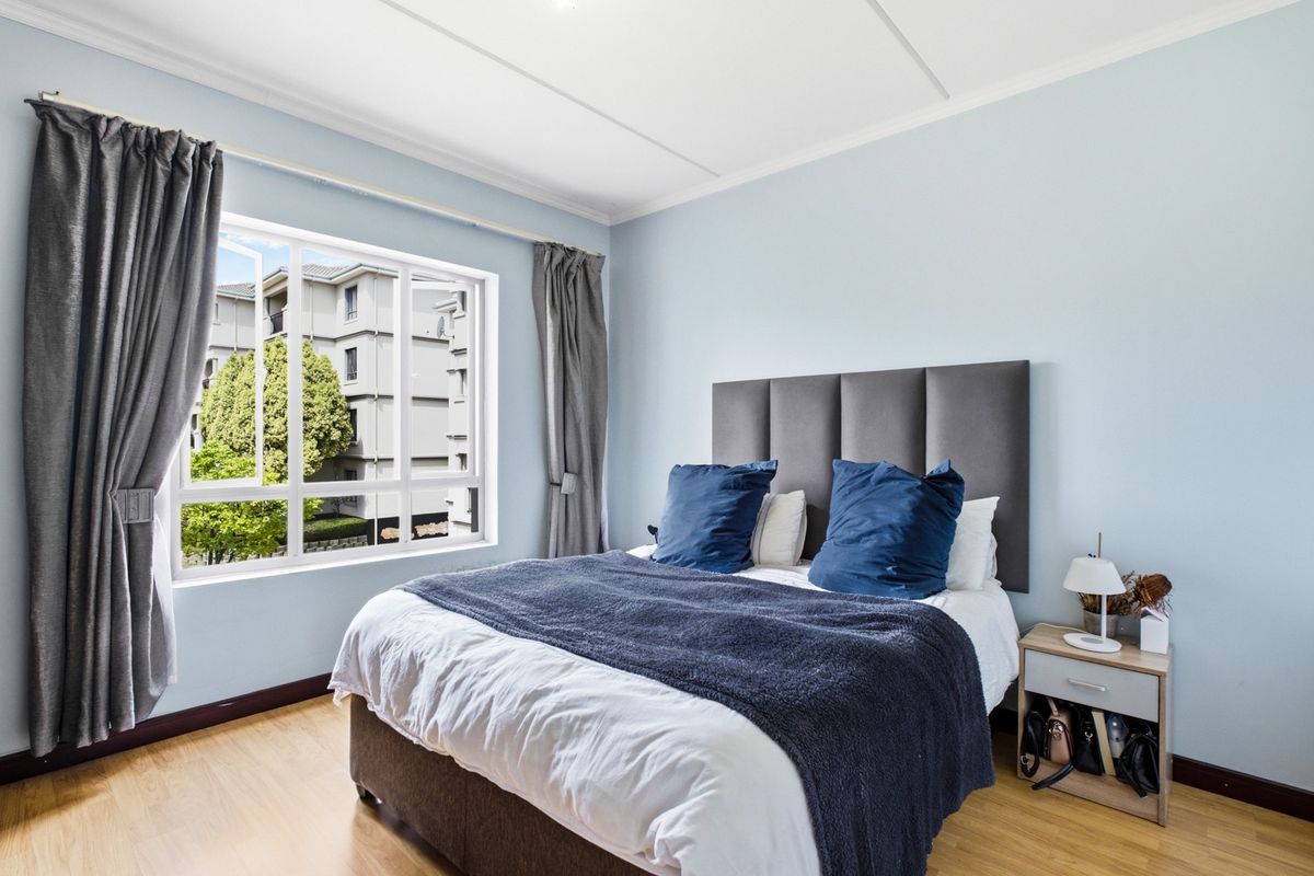 A well-lit bedroom featuring light-toned laminate flooring, neutral walls, and a large window providing natural light and an exterior view.