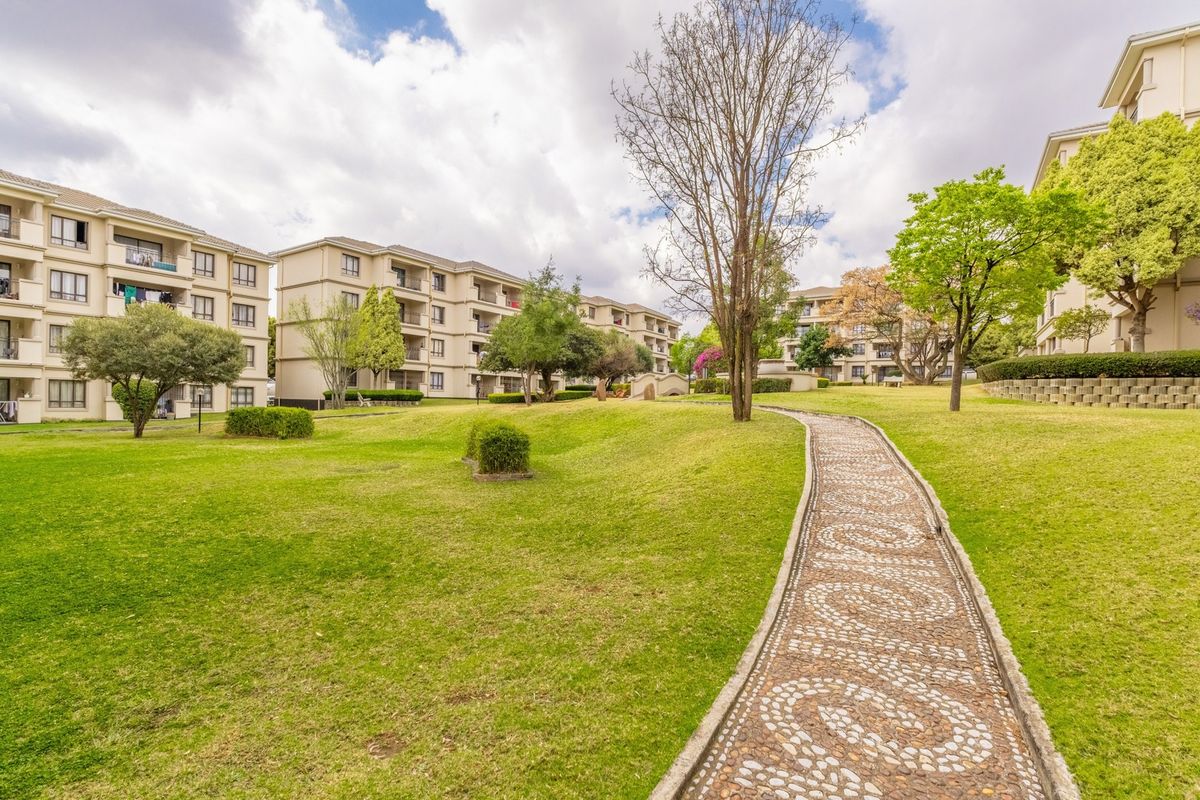 A well-maintained communal garden area featuring a decorative stone pathway, manicured lawns, and mature trees between multi-story apartment buildings.