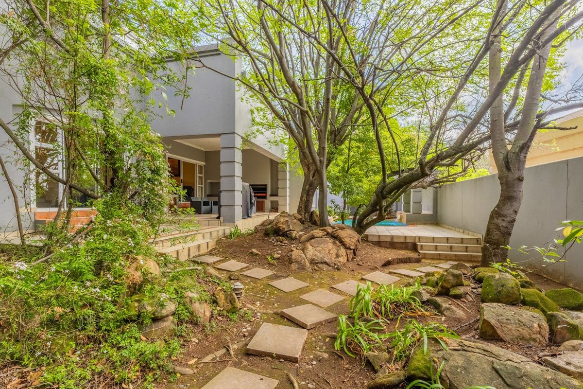 Rear garden area featuring a covered patio with built-in braai, a swimming pool, and a landscaped garden with mature trees and stone pathways.
