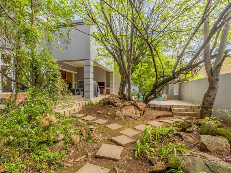 Rear garden area featuring a covered patio with built-in braai, a swimming pool, and a landscaped garden with mature trees and stone pathways.