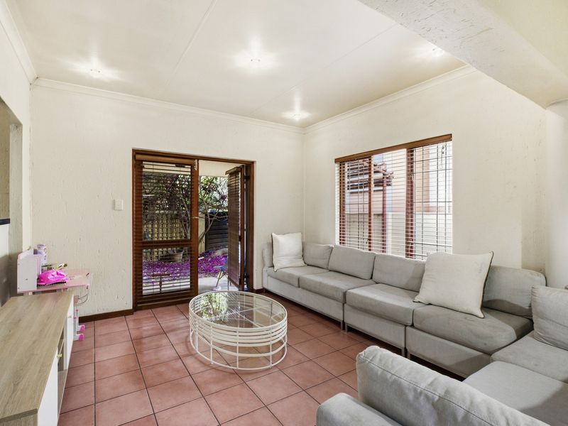A bright living room featuring tiled flooring, wooden framed windows with blinds, and direct access to a private outdoor area.