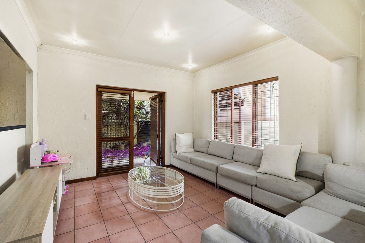 A bright living room featuring tiled flooring, wooden framed windows with blinds, and direct access to a private outdoor area.