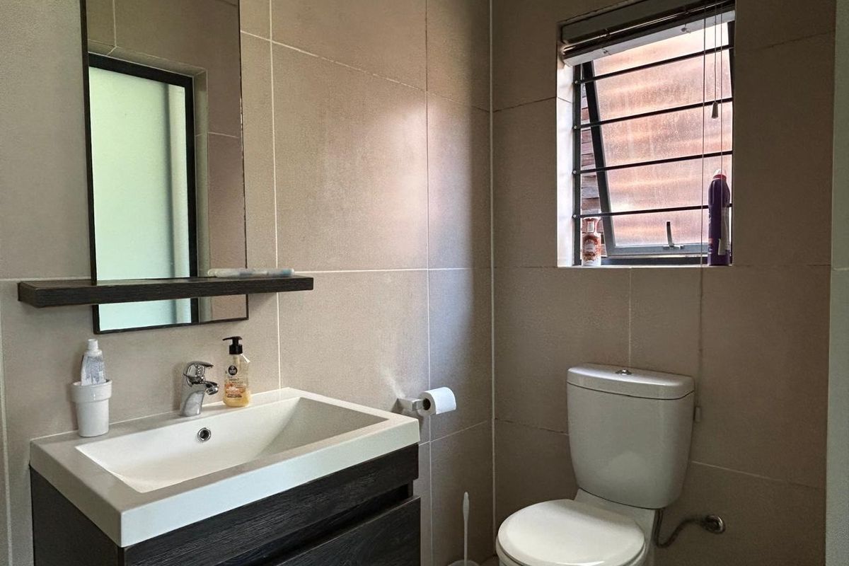 A modern bathroom featuring a vanity cabinet with basin, wall-mounted mirror, and a toilet, finished with neutral wall tiles and a window.