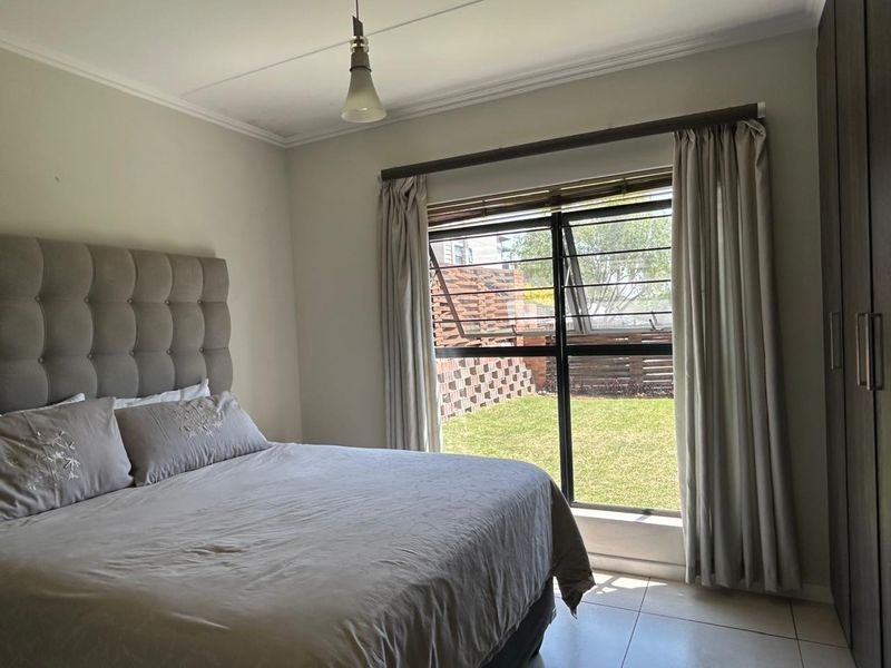 A well-lit bedroom featuring tiled flooring, a built-in wardrobe, and a large window with security bars overlooking a private garden area.