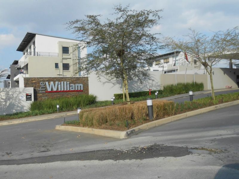The entrance facade of The William apartment complex, featuring stone-clad signage, perimeter security fencing, and a well-maintained paved driveway approach.