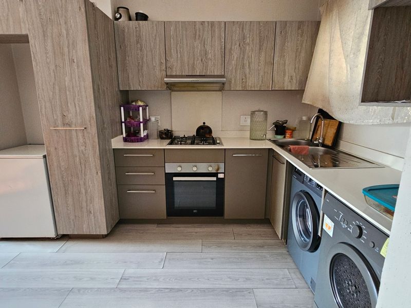 A functional kitchen featuring wood-look cabinetry, a gas hob, built-in oven, extractor fan, and integrated laundry appliances with modern laminate flooring.