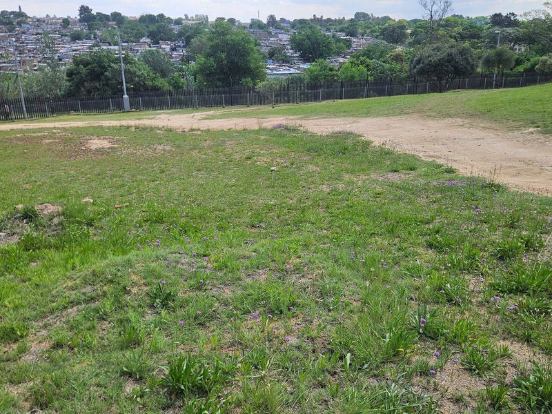 An elevated view overlooking a vacant residential plot with natural grass coverage and perimeter fencing, set against a suburban landscape.