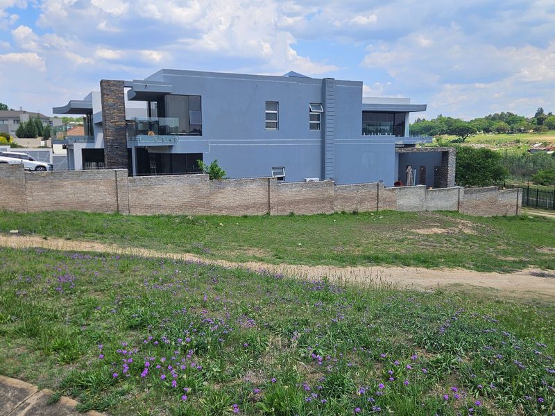 A view of a vacant residential plot featuring a brick boundary wall with a modern multi-story home visible in the background.