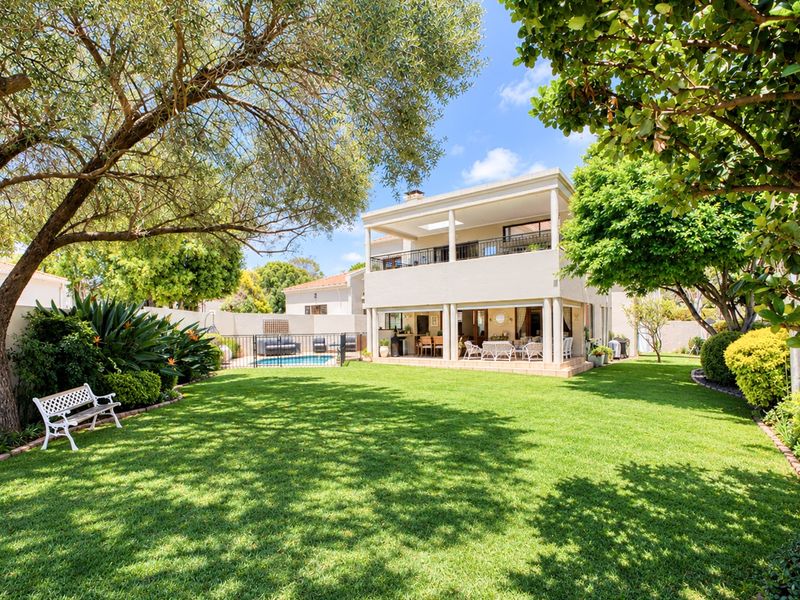 A spacious rear garden featuring a well-maintained lawn, mature trees, a swimming pool, and a two-story residence with a covered patio and upper balcony.