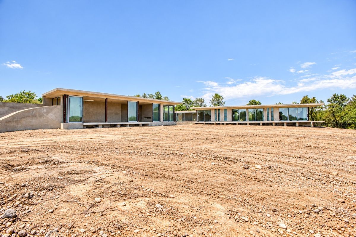 Modern architectural house exterior featuring extensive glass sliding doors, concrete structure, and cantilevered roof, set on a large, undeveloped plot of land.