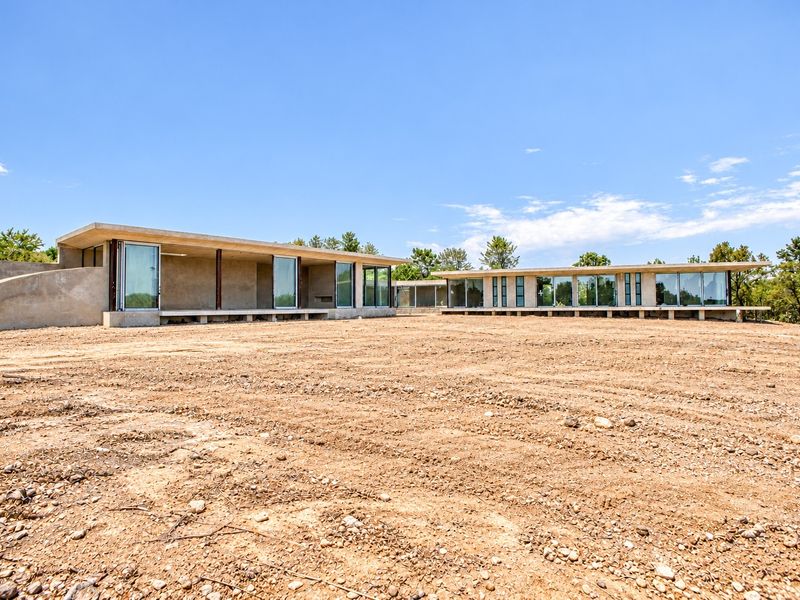 Modern architectural house exterior featuring extensive glass sliding doors, concrete structure, and cantilevered roof, set on a large, undeveloped plot of land.