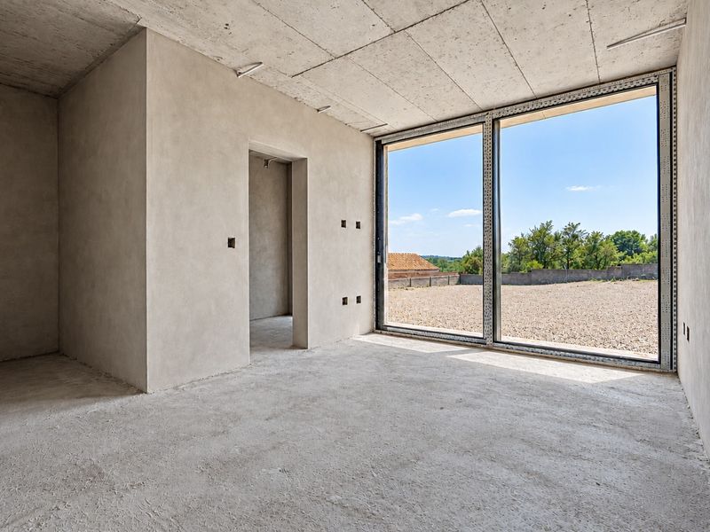 An unfinished interior space featuring large floor-to-ceiling windows, exposed concrete ceilings, and raw plastered walls, offering a blank canvas for custom finishes.
