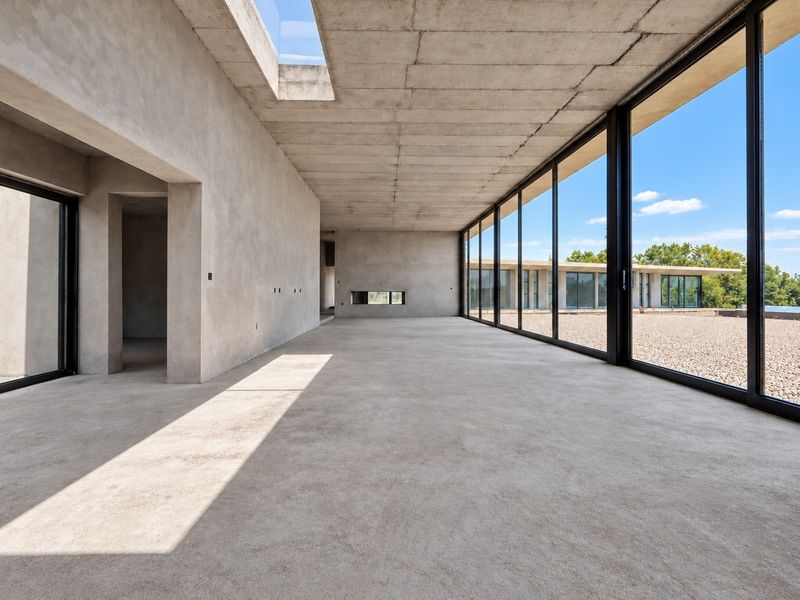 Spacious open-plan living area featuring raw concrete finishes, a large skylight, and expansive floor-to-ceiling glass doors opening to an outdoor gravel terrace.