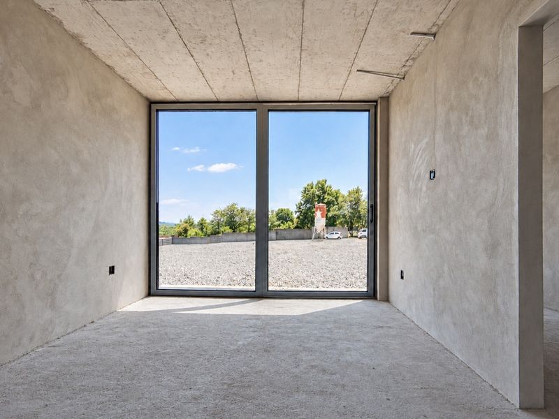 An unfinished interior space featuring raw concrete ceilings, plastered walls, and a large sliding glass door providing natural light and outdoor views.