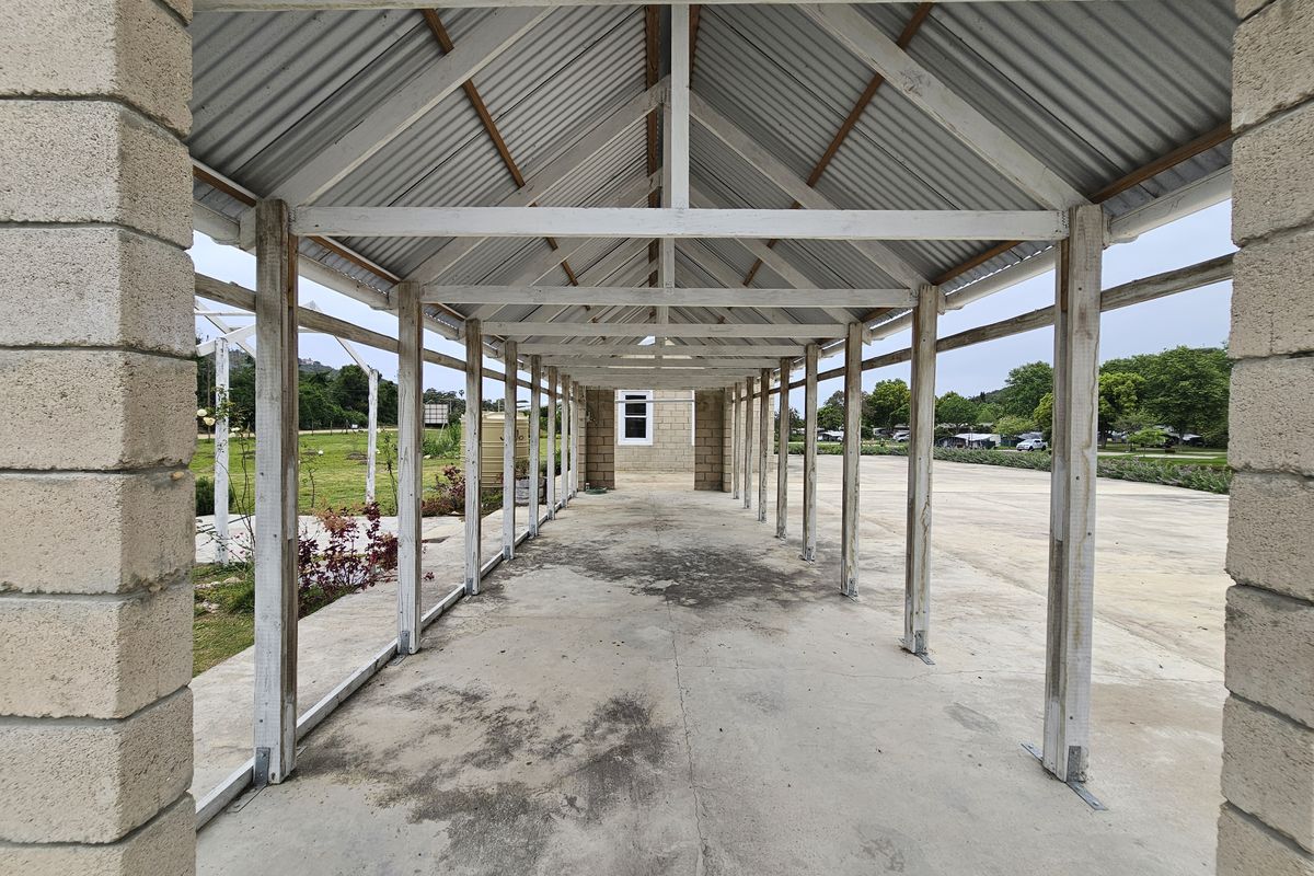 An open-air covered walkway or patio area featuring a timber frame structure with a corrugated metal roof and concrete flooring.