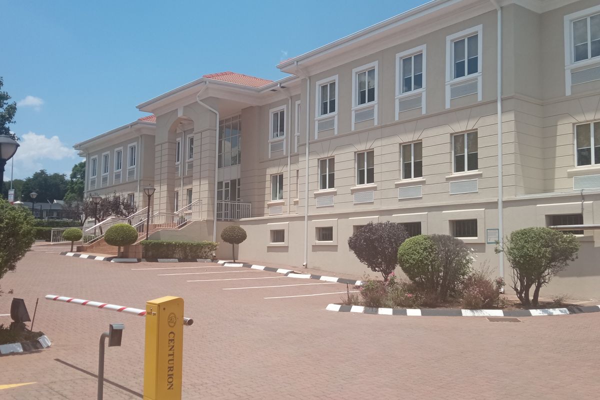 Exterior view of a multi-story office building featuring a paved parking area, security boom gate, and landscaped garden beds.