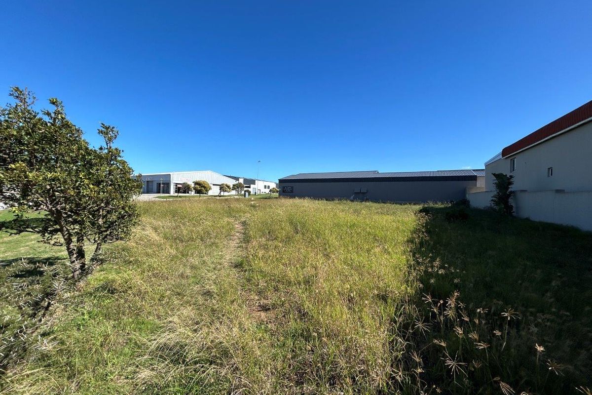 A view of a vacant commercial land plot featuring overgrown grass, situated in a developed commercial area with neighboring industrial buildings.