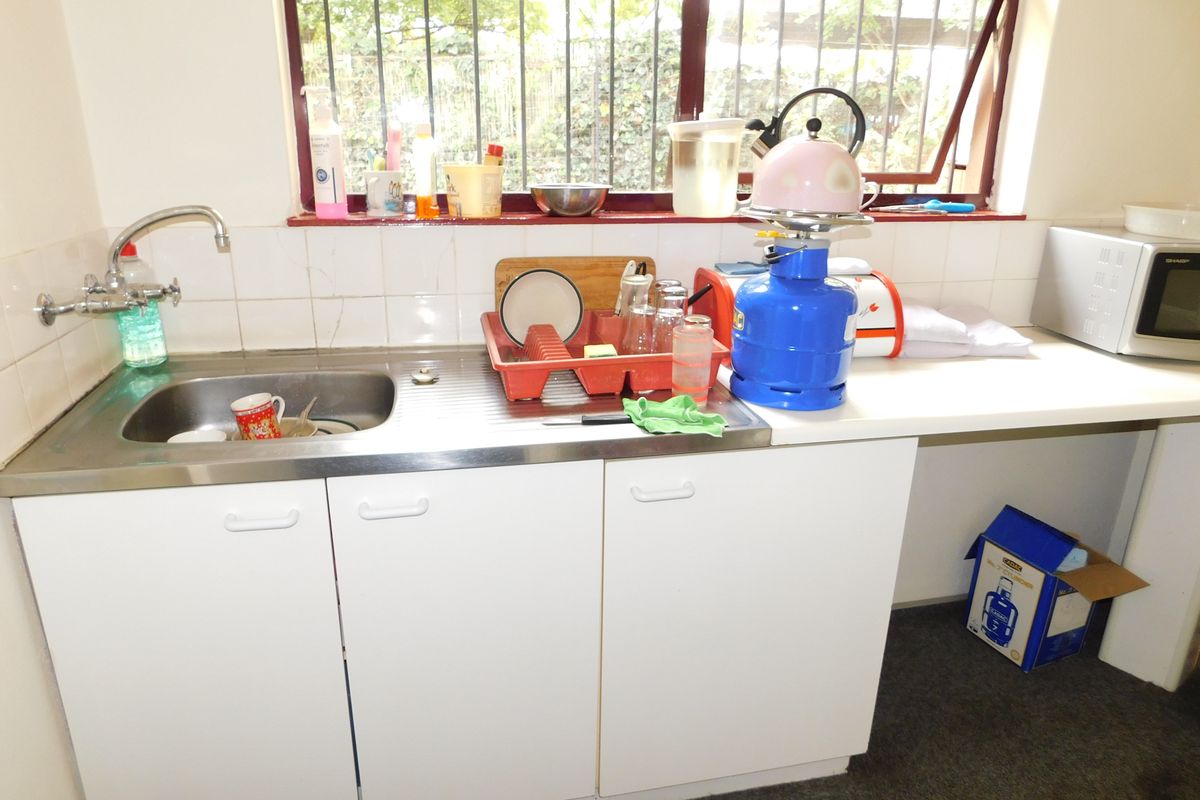 A functional kitchenette area featuring a stainless steel sink, white cabinetry, and tiled splashback, situated beneath a window with security bars.