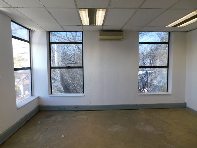 An empty office space featuring a suspended ceiling, recessed lighting, wall-mounted air conditioning, and large windows providing natural light.