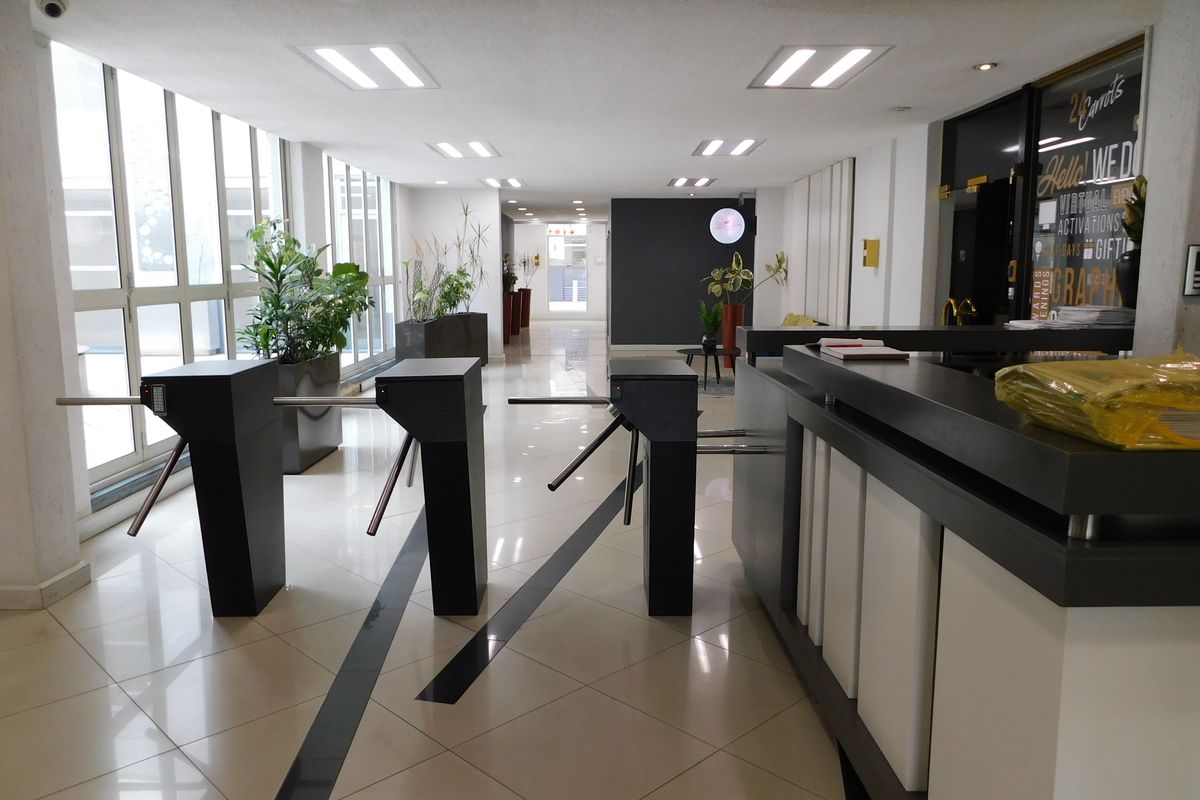Modern office building entrance featuring security turnstiles, a professional reception desk, and large windows providing ample natural light.