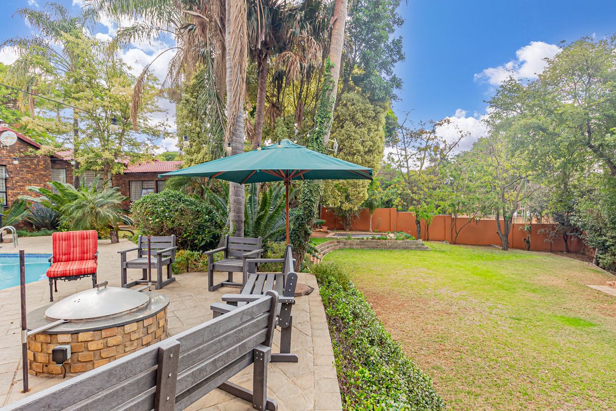 Spacious backyard featuring a paved patio area with a built-in fire pit, a swimming pool, and a well-maintained lawn surrounded by mature trees.