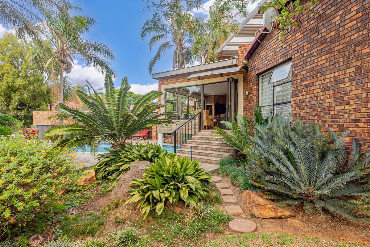 Rear view of the property featuring a brick facade, elevated patio access with stairs, and a landscaped garden area adjacent to the pool.
