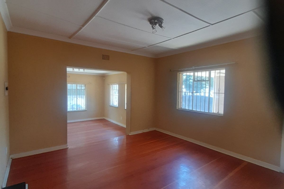 Spacious living area featuring polished wooden flooring, neutral wall tones, and large windows with security bars, leading into an adjacent room.