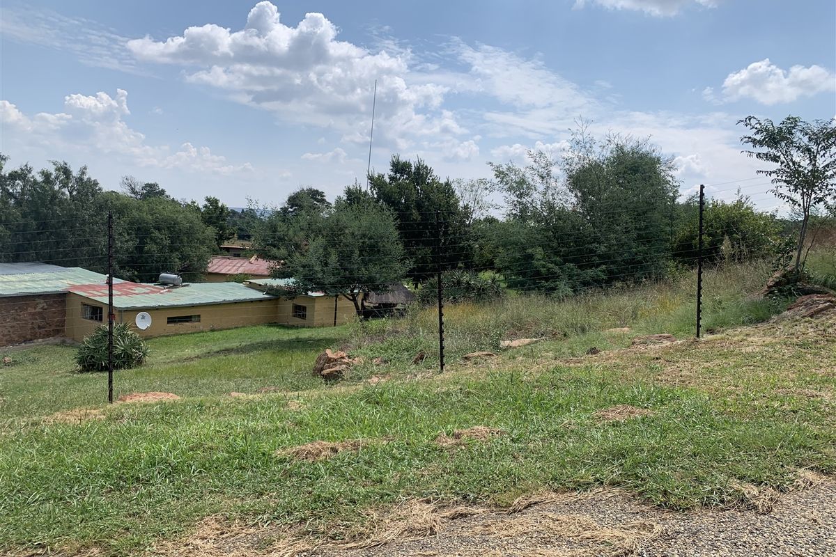 A view of a sloped residential plot featuring electric fencing and surrounding greenery, with neighboring structures visible in the background.