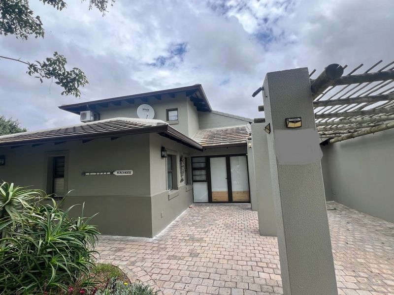 Front exterior view of the house featuring a paved entrance area, timber pergola, and modern neutral-toned facade with double glass door entry.