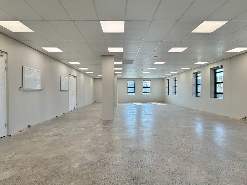 A spacious, open-plan commercial office area featuring polished concrete floors, a suspended ceiling with recessed lighting, and large windows providing natural light.