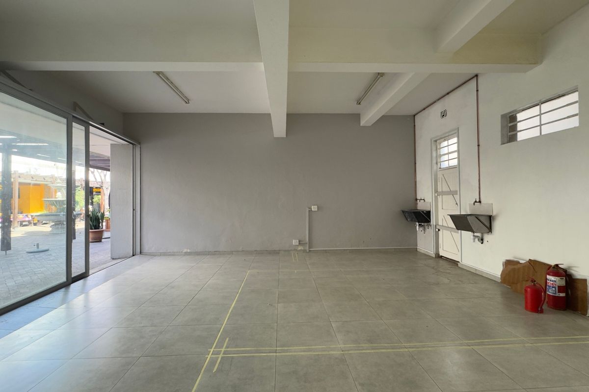 An empty open-plan commercial space featuring tiled flooring, exposed ceiling beams, large sliding glass doors, and wall-mounted stainless steel sinks.