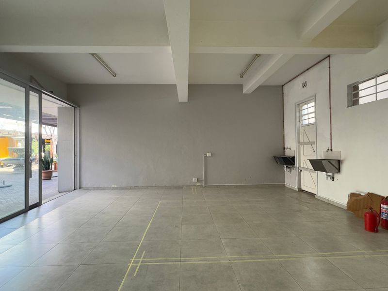 An empty open-plan commercial space featuring tiled flooring, exposed ceiling beams, large sliding glass doors, and wall-mounted stainless steel sinks.