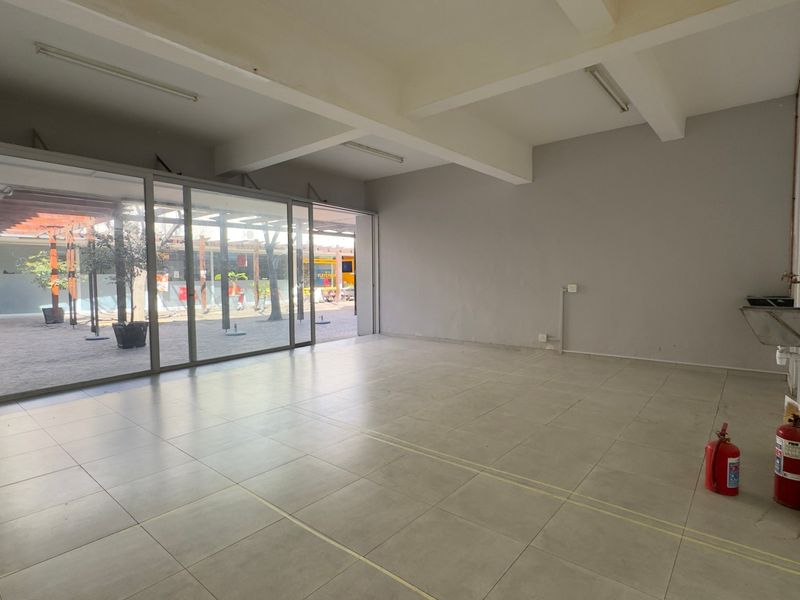 An empty open-plan commercial space featuring tiled flooring, exposed ceiling beams, and large sliding glass doors opening to an outdoor area.