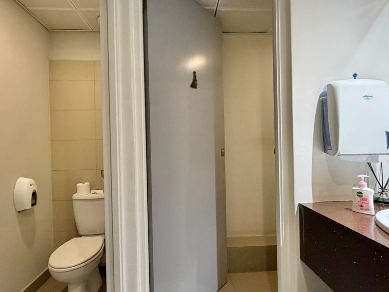 A functional office bathroom featuring a toilet, vanity with integrated washbasin, and wall-mounted utility dispensers.