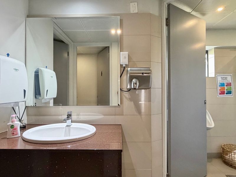 A functional office restroom featuring a vanity basin, wall-mounted mirror, hand dryer, paper towel dispenser, urinal, and a separate shower enclosure.