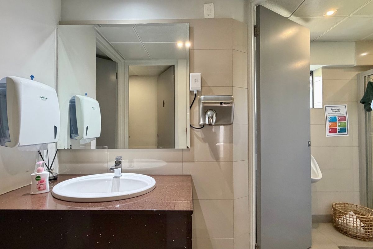 A functional office restroom featuring a vanity basin, wall-mounted mirror, hand dryer, paper towel dispenser, urinal, and a separate shower enclosure.