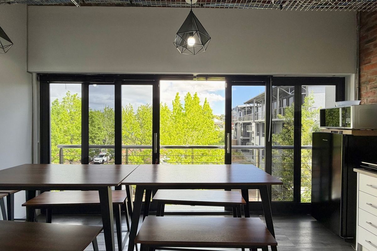 Modern open-plan office space featuring exposed brick accents, sleek black-framed sliding doors leading to a balcony, and contemporary pendant lighting.
