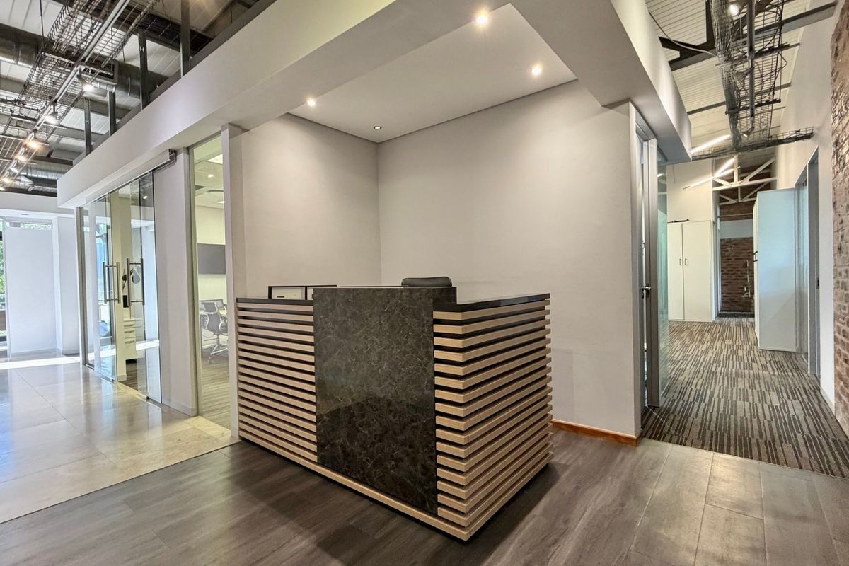Modern office reception area featuring a custom built-in desk, exposed industrial ceiling elements, glass partitions, and a combination of laminate and tiled flooring.
