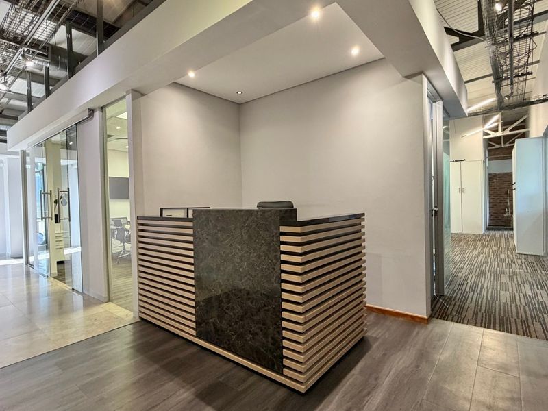 Modern office reception area featuring a custom built-in desk, exposed industrial ceiling elements, glass partitions, and a combination of laminate and tiled flooring.