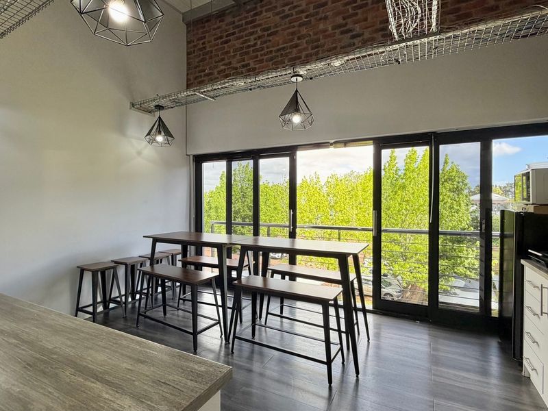 Modern open-plan office space featuring exposed brickwork, industrial-style cable trays, pendant lighting, and large sliding glass doors leading to an outdoor balcony.