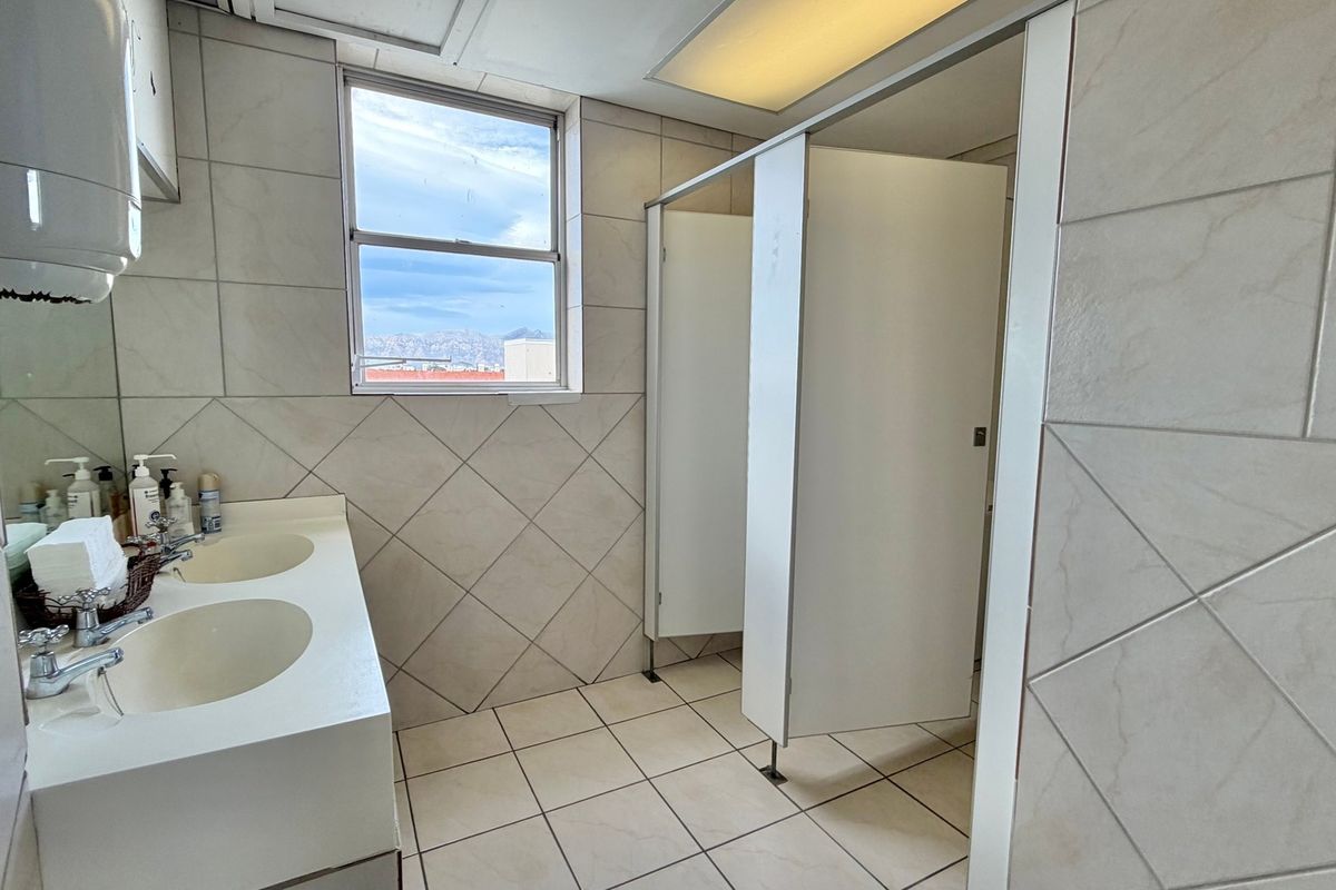 A functional office bathroom featuring a dual vanity basin, tiled surfaces, and multiple toilet cubicles with a view of the surrounding area.