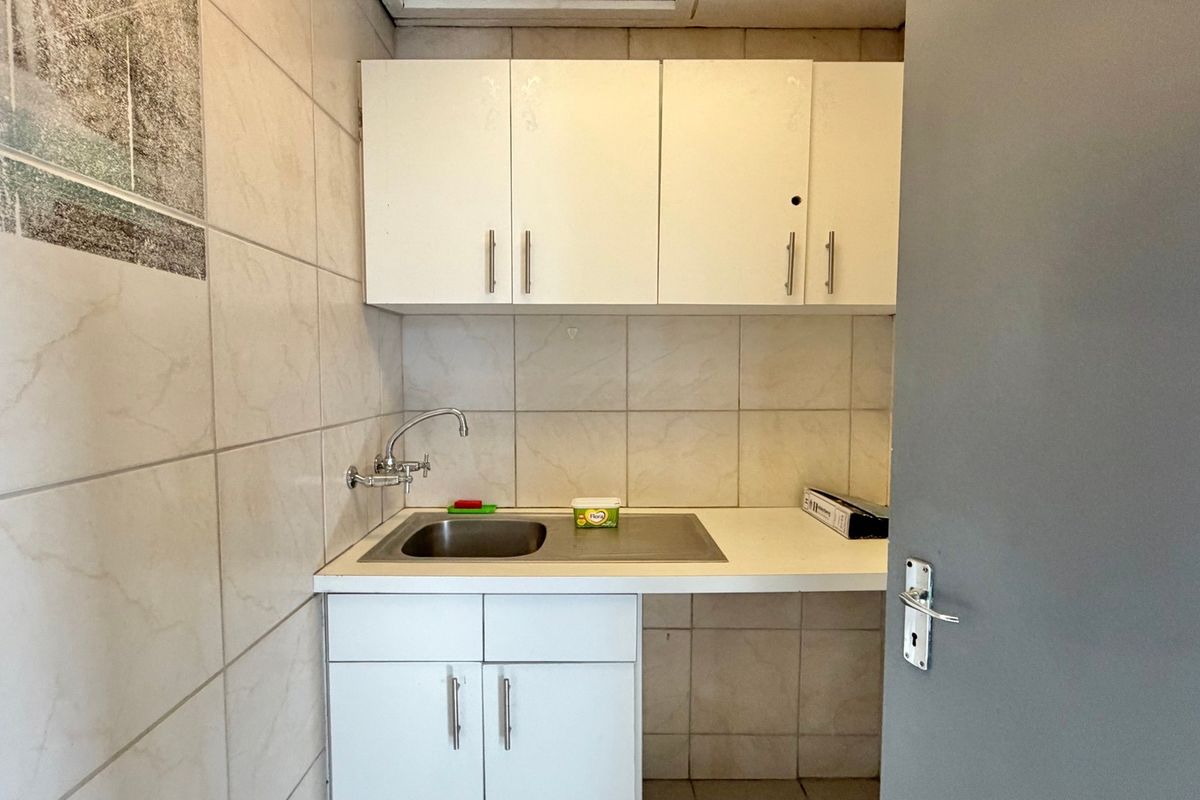 A compact office kitchenette featuring white built-in cabinetry, a stainless steel sink, and tiled wall finishes.