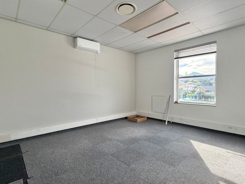 A vacant office space featuring a suspended ceiling, wall-mounted air conditioning, carpeted flooring, and a window with natural light.