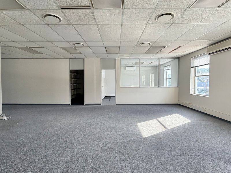 A spacious, empty office area featuring a suspended ceiling, carpeted flooring, split-system air conditioning, and a glass partition wall for a professional layout.
