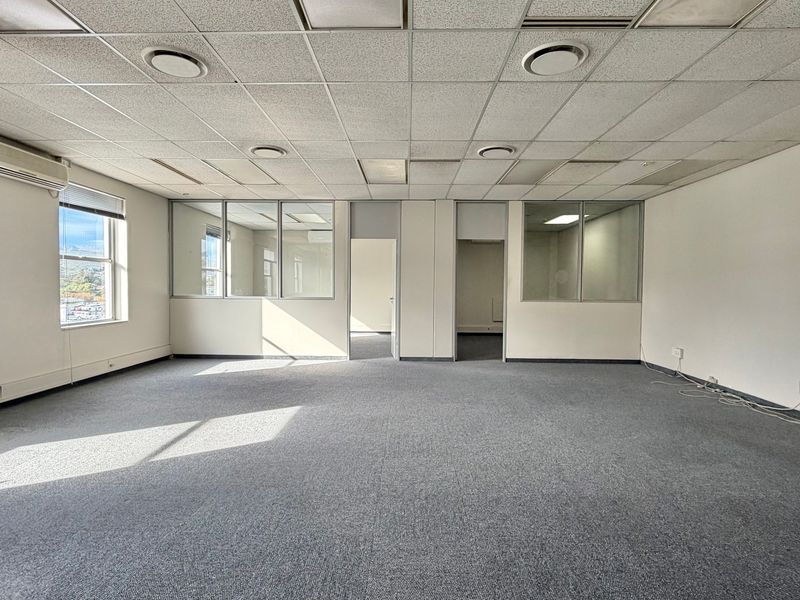 An empty, open-plan office space featuring glass partitions, carpeted flooring, a suspended ceiling with recessed lighting, and wall-mounted air conditioning units.