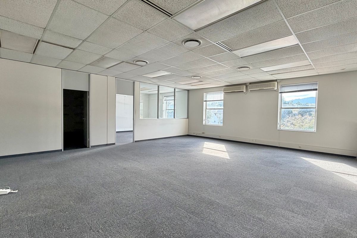 A spacious, vacant office area featuring a suspended ceiling, recessed lighting, split-system air conditioning, and large windows providing natural light.