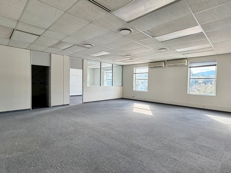 A spacious, vacant office area featuring a suspended ceiling, recessed lighting, split-system air conditioning, and large windows providing natural light.