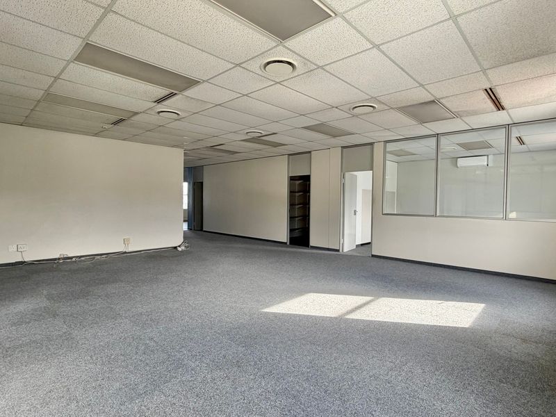 Spacious, empty open-plan office area featuring suspended ceilings, recessed lighting, air conditioning, and glass partition walls with grey carpeting.