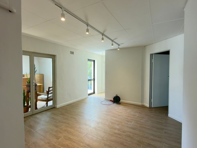 A bright, open-plan office space featuring modern wood-look tiled flooring, track lighting, and neutral white walls, offering a clean and versatile workspace.