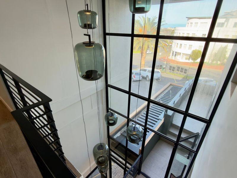 A modern, double-volume hallway featuring floor-to-ceiling steel-framed windows, elegant pendant lighting, and a sleek metal staircase railing overlooking the exterior.